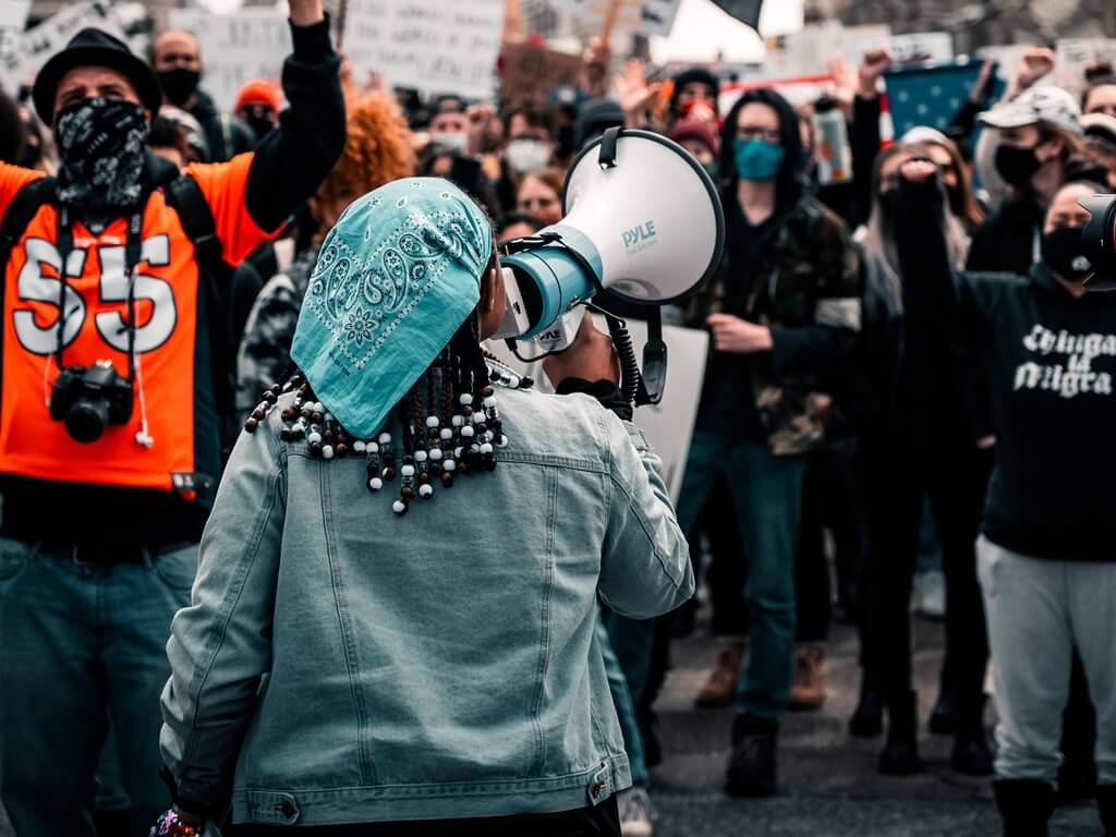A crowd of people involved in protest action. A young woman addresses crowd through loudhaler.
